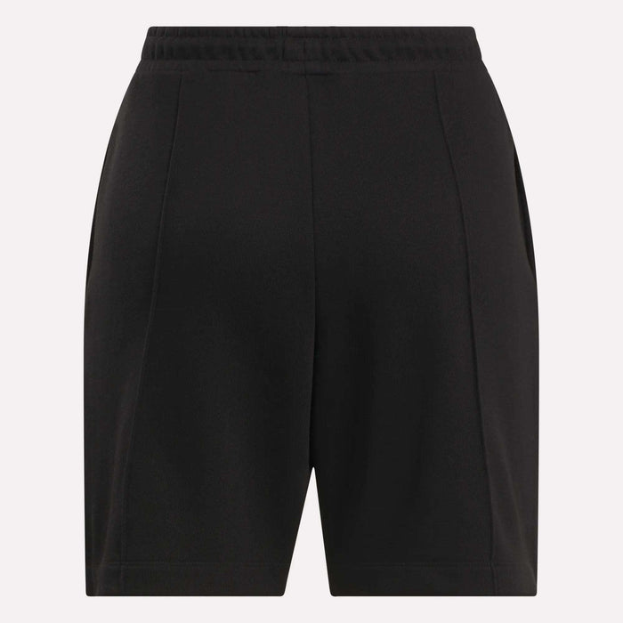 Reebok Women's Pintuck Long Shorts in black, featuring an elastic waistband, front pleats, and side pockets, made from a soft cotton-blend fleece. Displayed against a plain light background.