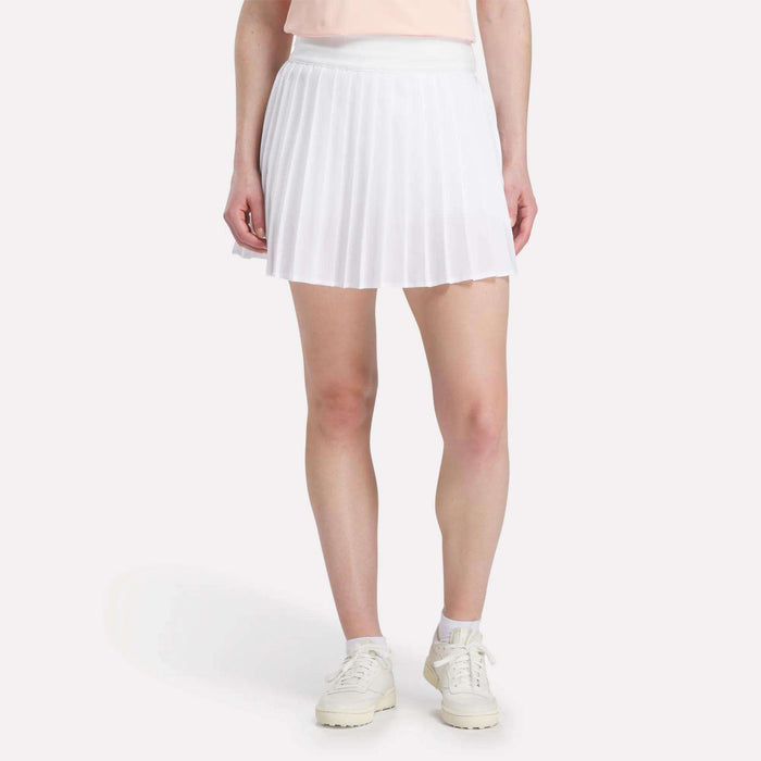 A woman wears a white Reebok Women's Pleated Skort with white sneakers and a light pink top, standing against a plain light background. The cropped photo shows her outfit from the waist down.