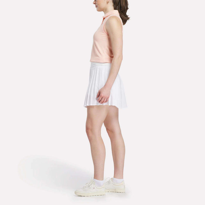 A woman stands in profile against a white background, wearing a sleeveless light pink top, Reebok Women's Pleated Skort, white socks, and white sneakers.