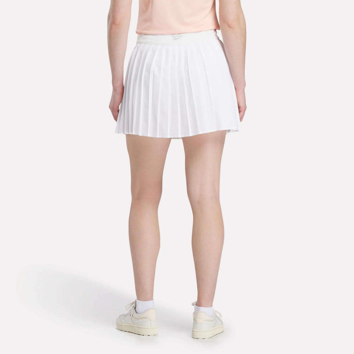 A person faces away, wearing a light pink shirt and the Reebok Women's Pleated Skort, paired with white socks and white sneakers against a plain white background.