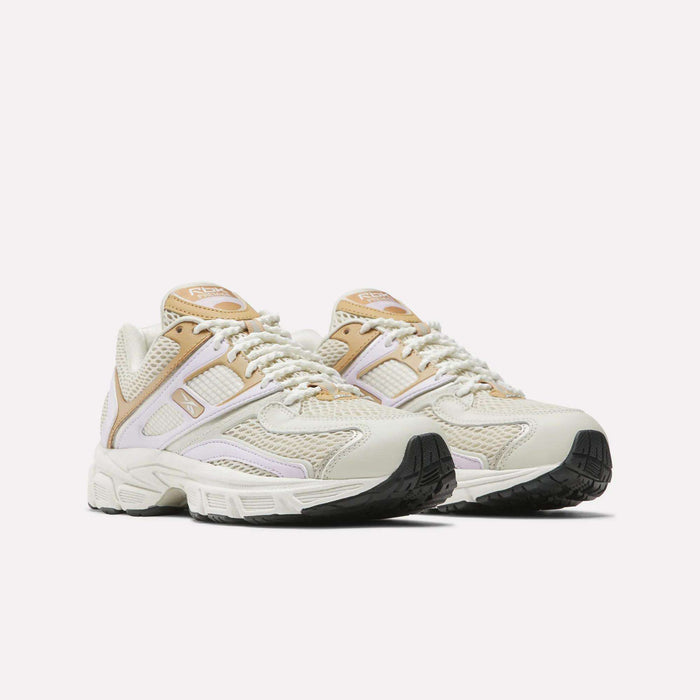 The Reebok Women's Premier Trinity Shoes feature beige and white uppers with tan accents, mesh panels, and chunky black soles, blending retro tech style with modern comfort. Displayed on a plain white background.