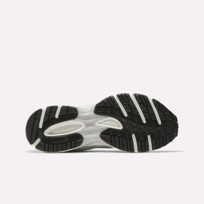 The image shows the bottom view of Reebok Women's Premier Trinity Shoes, highlighting a black and white rubber outsole with textured tread for traction. The retro tech design is displayed on a plain white background.