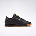 The Reebok Women's Princess Shoes in black feature a gum sole, black laces, and gold Reebok branding on the side, tongue, and heel. Combining cushioned comfort with heritage style, they're displayed against a plain light gray background.