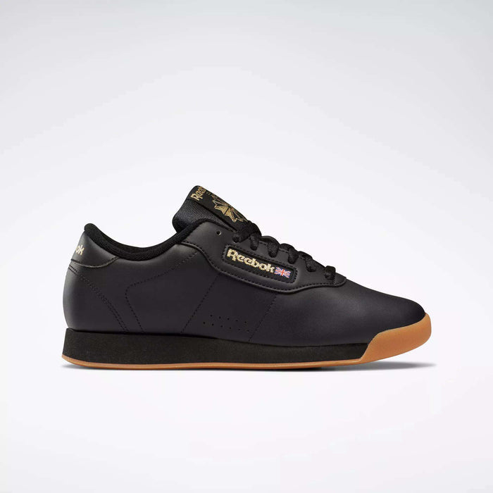 The Reebok Women's Princess Shoes in black feature a gum sole, black laces, and gold Reebok branding on the side, tongue, and heel. Combining cushioned comfort with heritage style, they're displayed against a plain light gray background.