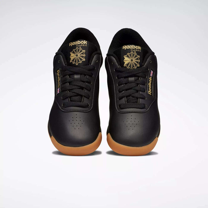 A pair of Reebok Women's Princess Shoes in black with gum soles and gold logos, featuring a cushioned design, are placed side by side on a plain light background, facing forward.