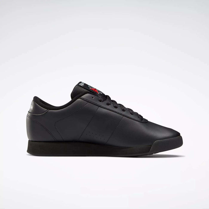 The Reebok Women's Princess Shoes are black low-top sneakers with a sleek, simple design, cushioned comfort, black laces, and subtle Reebok branding on the tongue and heel, displayed in a side profile on a light gray background.