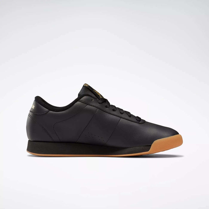 The Reebok Women's Princess Shoes are classic black low-tops with black laces, cushioned comfort, a gum sole, and subtle gold branding, showcased against a plain light background.