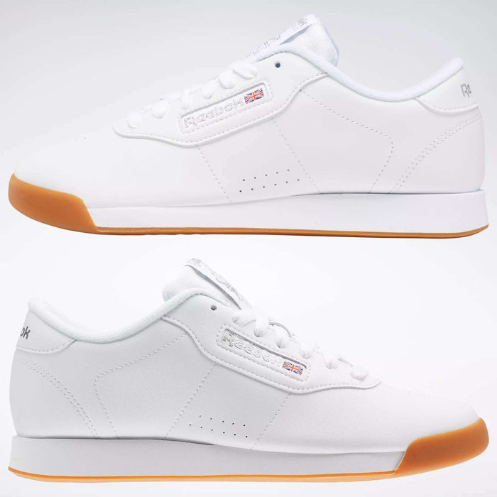 Two Reebok Women's Princess Shoes in white with gum soles sit side by side, facing opposite ways on a light background. Featuring simple stitching, soft cushioning, and the Reebok logo with a small British flag on the side.