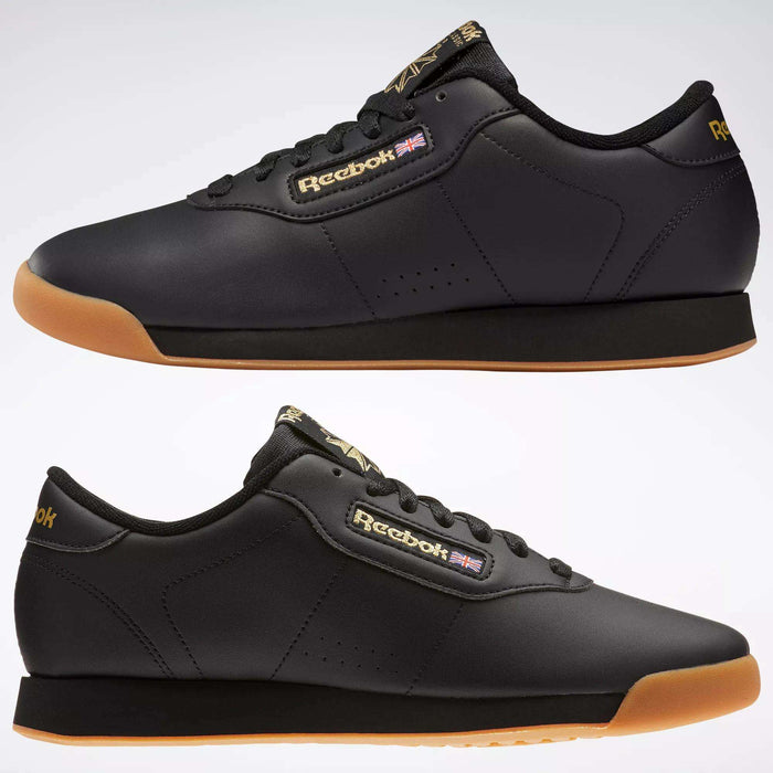 Two black Reebok Women's Princess Shoes with gum soles are shown from the side on a light background. Featuring the Reebok logo and Union Jack, these sneakers blend heritage style with modern comfort.