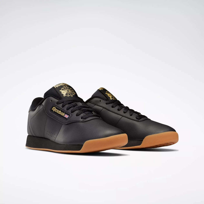 Reebok Women's Princess Shoes, in black with gum soles and gold logo accents, offer classic heritage style on a light gray background. These cushioned sneakers deliver comfort with every step.
