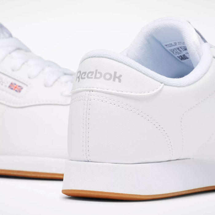 Close-up of Reebok Women's Princess Shoes in white with gum soles, featuring visible Reebok branding and a small Union Jack flag. These classic sneakers offer cushioned insoles for comfort and a clean, minimalist design.