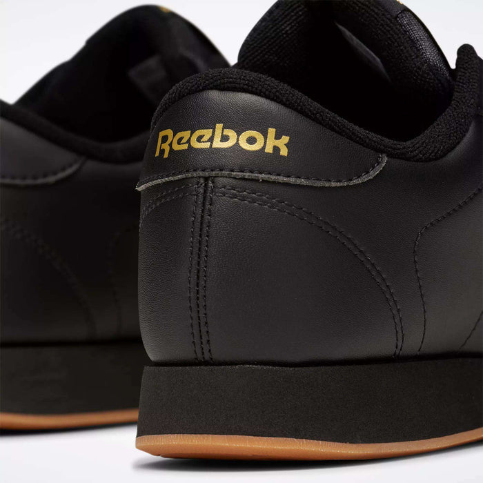 Close-up of the back of two black Reebok Women's Princess Shoes with gold branding on the heel and gum-colored rubber outsoles, highlighting their heritage style against a white background.