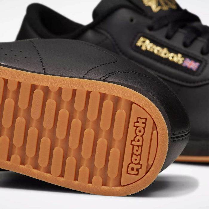 Close-up of black Reebok Women's Princess Shoes with gum soles, highlighting the textured sole pattern and Reebok branding. The heritage-inspired design features the Reebok logo and Union Jack flag on the side of this classic women's sneaker.