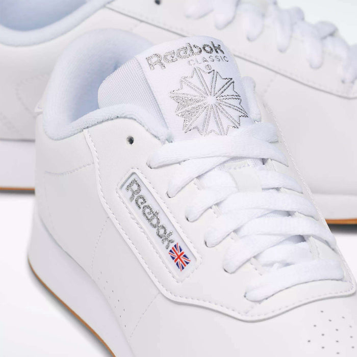 Close-up of Reebok Women's Princess Shoes, featuring a classic white design with the Reebok logo, signature Union Jack flag, star detail on the tongue, visible cushioned accents, and coordinating white laces and stitching.