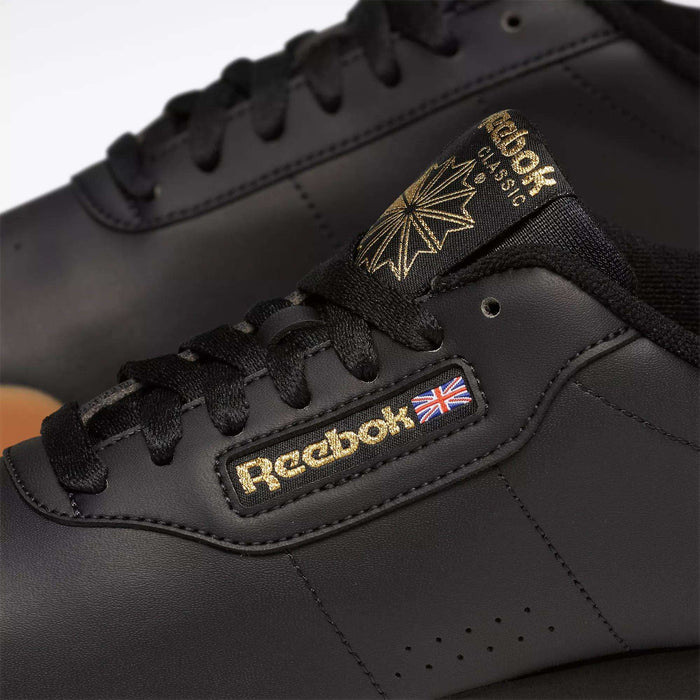 Close-up of black Reebok Women's Princess Shoes featuring gold Reebok branding, a British flag patch on the side, and black laces—a heritage style women's classic shoe with timeless appeal.