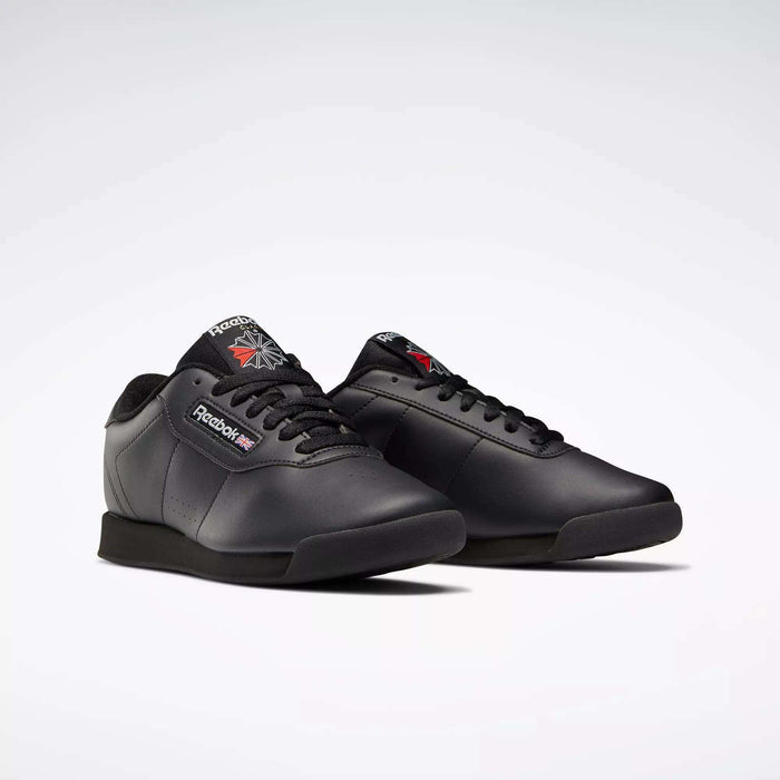 A pair of black Reebok Women's Princess Wide Shoes with black soles and laces, featuring the classic design and the Union Jack logo on the side and tongue, displayed on a plain light background.