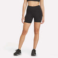 A woman wearing a black sports bra, Women's Reebok x F45 Training High-Rise Biker Shorts, and black running shoes stands against a plain white background.