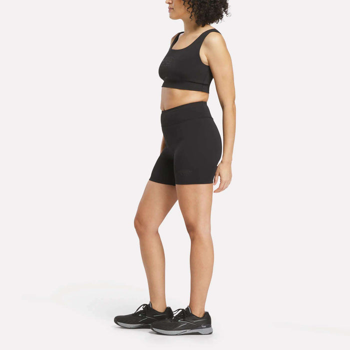 A woman stands against a plain white background, facing sideways with her arms relaxed, wearing a black Reebok performance sports bra, Women's Reebok x F45 Training High-Rise Biker Shorts in black, and black running shoes.
