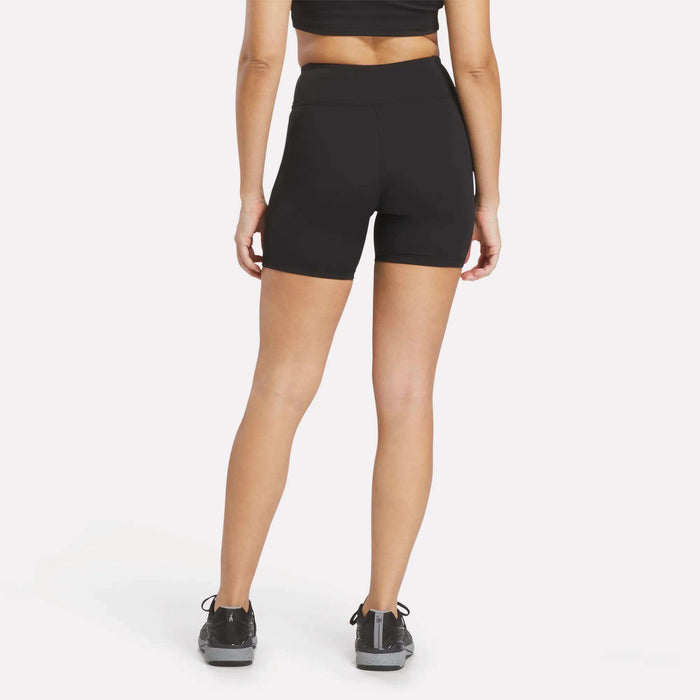 A woman stands facing away, wearing a black Reebok performance sports bra, Women's Reebok x F45 Training High-Rise Biker Shorts in black, and black sneakers. The background is plain white.