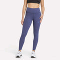 A person stands against a plain white background wearing a black sports bra, Women's Reebok x F45 Training Leggings in Legacy Blue, and white sneakers.