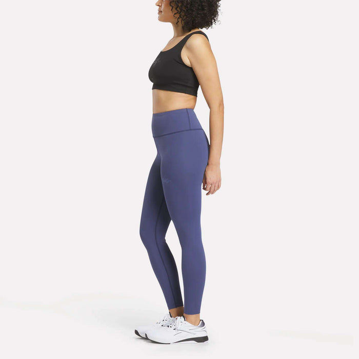 A woman stands in profile against a plain white background, wearing a black sports bra, crisp white sneakers, and the Women's Reebok x F45 Training Leggings—a supportive, high-waisted blue style from Reebok—perfect for an intense F45 Training workout.