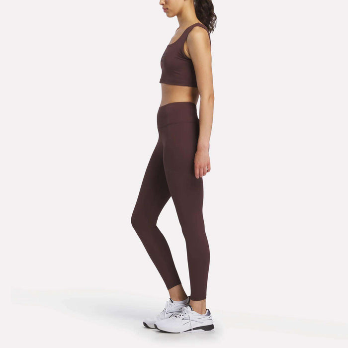 A woman stands in profile against a plain white background, wearing Women's Reebok x F45 Training Leggings and a burgundy sports bra, paired with white Reebok athletic shoes.