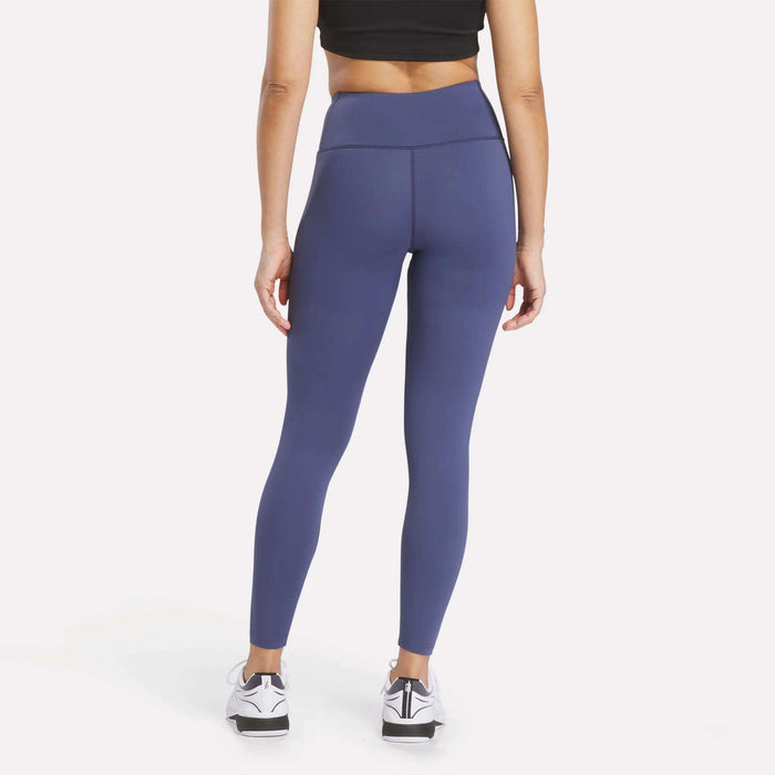A person stands facing away, wearing a black crop top paired with high-waisted blue Women's Reebok x F45 Training Leggings and white sneakers. The sporty ensemble by Reebok is ideal for F45 Training, set against a plain, light-colored background.