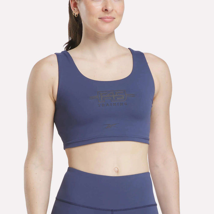 A woman wearing a Legacy Blue Women's Reebok x F45 Training Sports Bra stands against a plain light background, facing forward and smiling slightly.