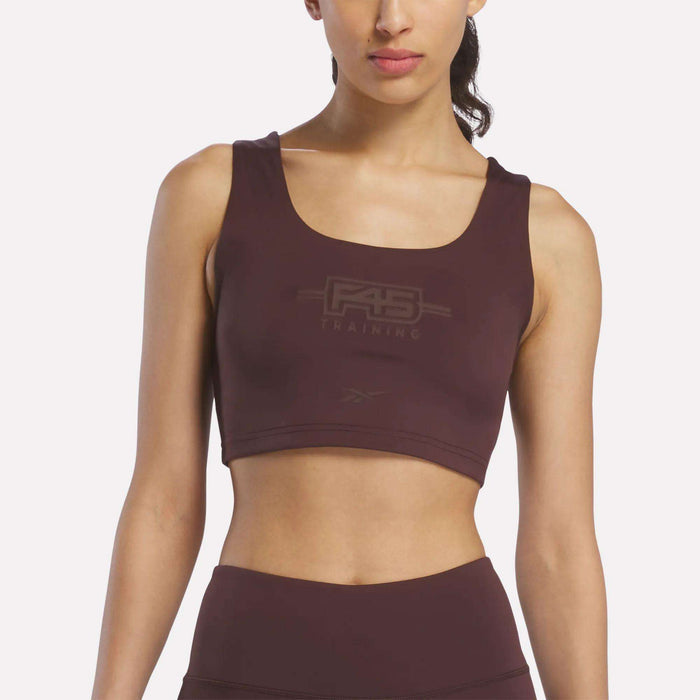A woman wearing a Reebok Women's Reebok x F45 Training Sports Bra in Dark Cocoa stands against a plain white background, facing forward with her head slightly turned.