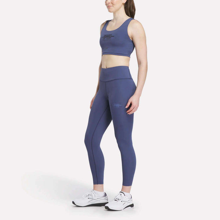 Outfitted in the Women's Reebok x F45 Training Sports Bra, a woman stands and smiles against a plain white background, paired with matching blue leggings and white athletic shoes—ready for her next F45 Training session.