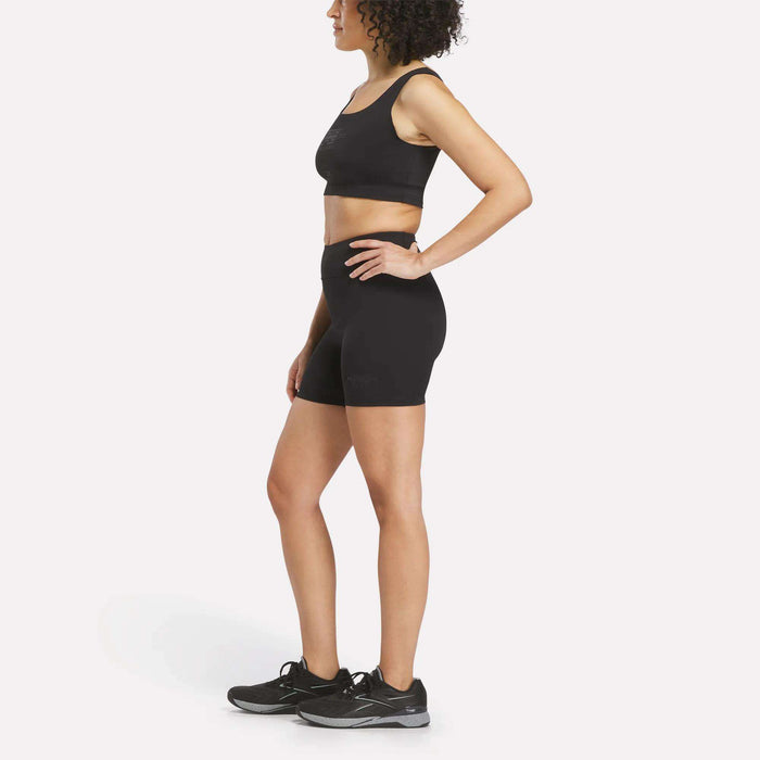 A woman stands in profile against a plain white background, wearing a black Women's Reebok x F45 Training Sports Bra, paired with black high-waisted biker shorts and black running shoes.