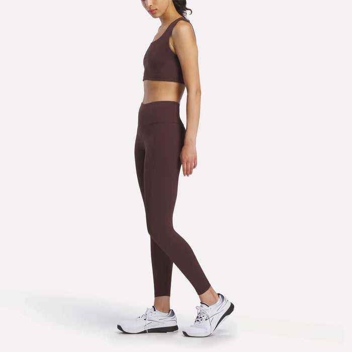 A woman stands sideways against a plain white background, wearing brown athletic leggings and the Women's Reebok x F45 Training Sports Bra by Reebok, paired with white sneakers.