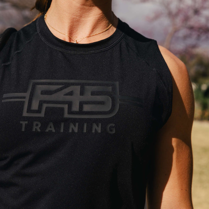 Close-up of a woman wearing the Women's Reebok x F45 Training Tank Top, a black sweat-wicking piece by Reebok featuring the F45 Training logo, set against a sunlit outdoor backdrop that suggests intense exercise among trees.