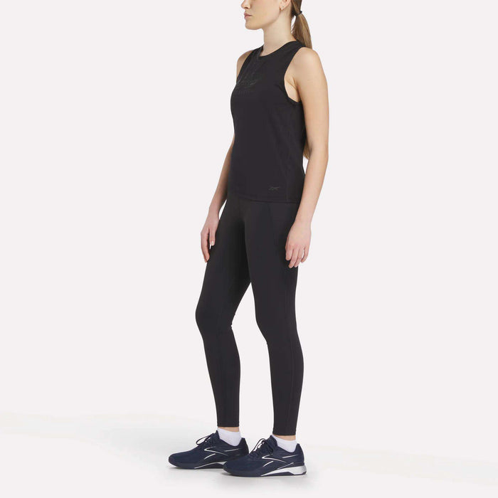 A woman with a ponytail stands slightly to the side wearing the Reebok x F45 Training Tank Top in black, paired with black leggings and navy running shoes. The sweat-wicking Reebok outfit pops against the plain white background.