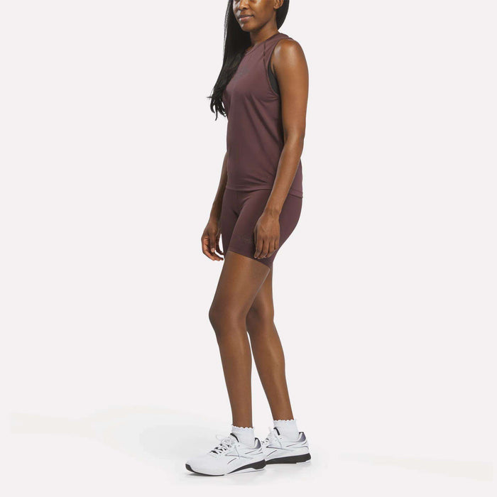 A woman wearing a Women's Reebok x F45 Training Tank Top as part of her sleeveless maroon sports outfit and white athletic shoes stands against a plain white background, facing slightly to the left.