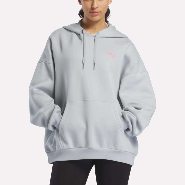 A person wearing the Reebok Women's Relaxed Fleece Hoodie in Breeze—a light gray oversized design with a front pocket and drawstrings—stands with hands tucked in the pocket. This cozy fleece hoodie is finished with a small pink logo on the left chest for a simple yet stylish touch.