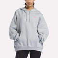 A person wearing the Reebok Women's Relaxed Fleece Hoodie in Breeze—a light gray oversized design with a front pocket and drawstrings—stands with hands tucked in the pocket. This cozy fleece hoodie is finished with a small pink logo on the left chest for a simple yet stylish touch.