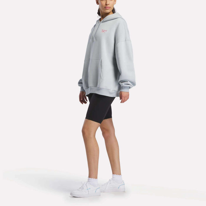 A person wearing a Reebok Women's Relaxed Fleece Hoodie in light gray, styled with black bike shorts, white socks, and white sneakers stands against a plain white background.