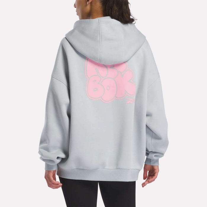 A woman models the Reebok Women's Relaxed Fleece Hoodie in light gray, featuring an oversized fit and a pink graffiti-style Reebok logo on the back, standing against a plain white background.