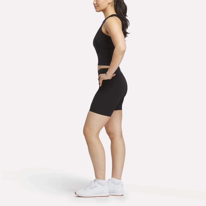A woman wearing a Reebok Women's Rib Tank Top in black, paired with black fitted shorts and white athletic shoes, stands in profile against a plain white background.
