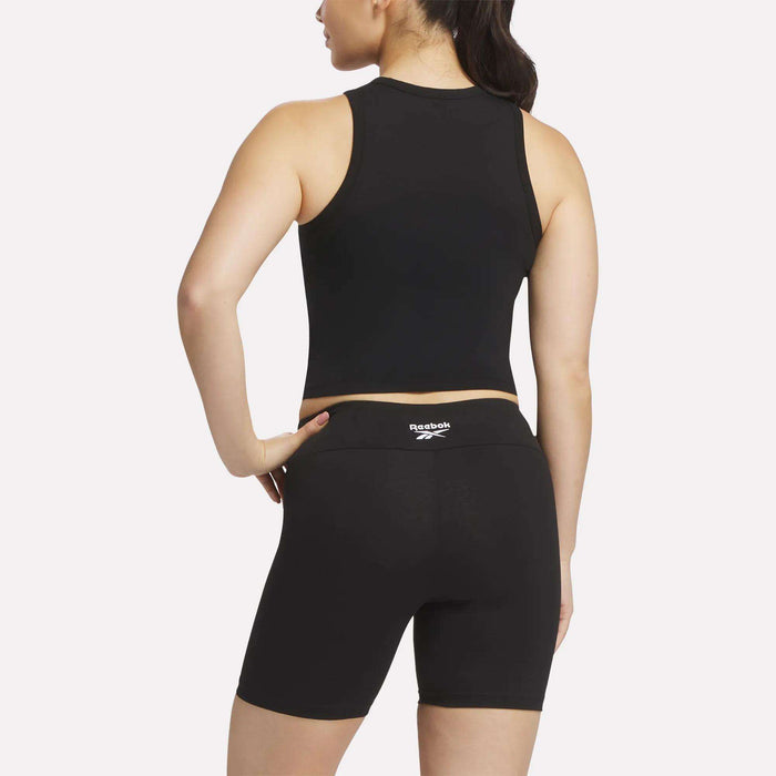 A woman stands with her back to the camera, wearing a black Reebok Women's Rib Tank Top and black biker shorts. Her right hand rests on her hip, and her hair is tied back.