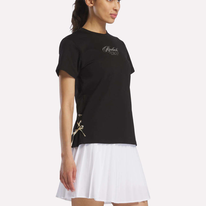 A woman wears the Reebok Women's Ribbon T-Shirt in black, crafted from soft cotton and featuring a small logo text on the chest, paired with a white pleated skirt and standing against a plain light background.
