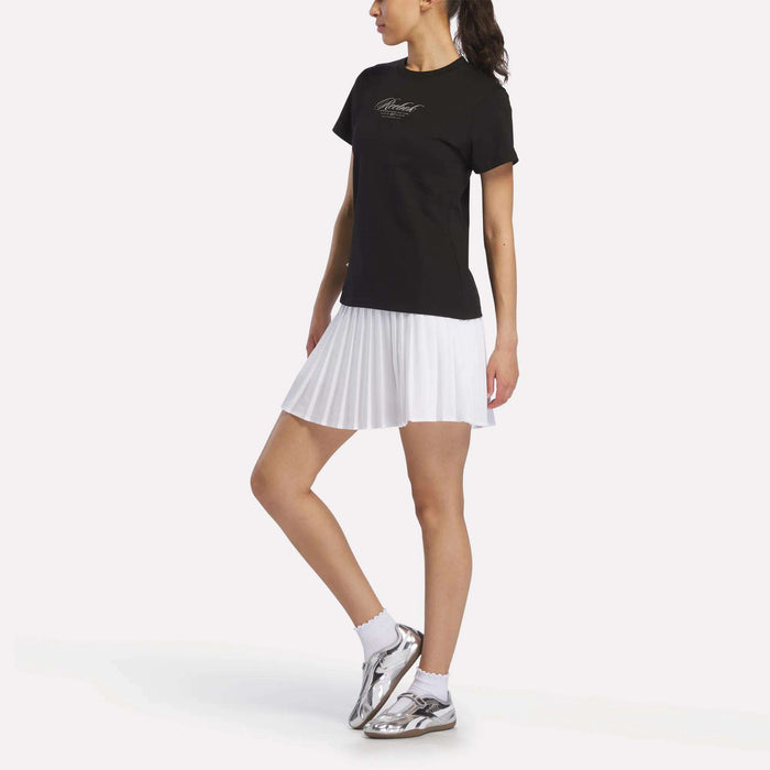 A woman wearing a Reebok Women's Ribbon T-Shirt in soft black cotton, paired with a white pleated skirt, white socks, and silver sneakers stands with one leg slightly bent, looking to the side against a plain white background.