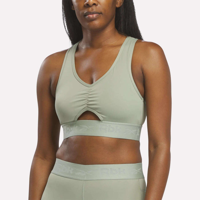 A woman wearing the Reebok Women's Ruched Sports Bra in Hazy Green, featuring a front keyhole cutout and matching leggings for medium-impact performance, stands against a plain background.