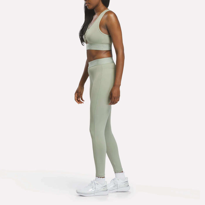 A woman stands sideways against a plain white background, wearing the Reebok Women's Ruched Sports Bra in sage green with matching leggings and white sneakers. This medium-impact sports bra is designed with advanced performance technology by Reebok, delivering style and support for active lifestyles.