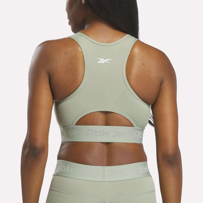 A woman is shown from the back wearing the Reebok Women's Ruched Sports Bra in light green, highlighting its racerback design, small cutout detail, and medium-impact support.