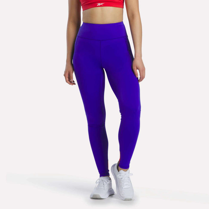 A person is seen wearing a red sports bra, white sneakers, and Reebok Women's Running Leggings in Breakout Blue/Vector Red, featuring mesh panels. They are standing against a plain white background with their face not visible.