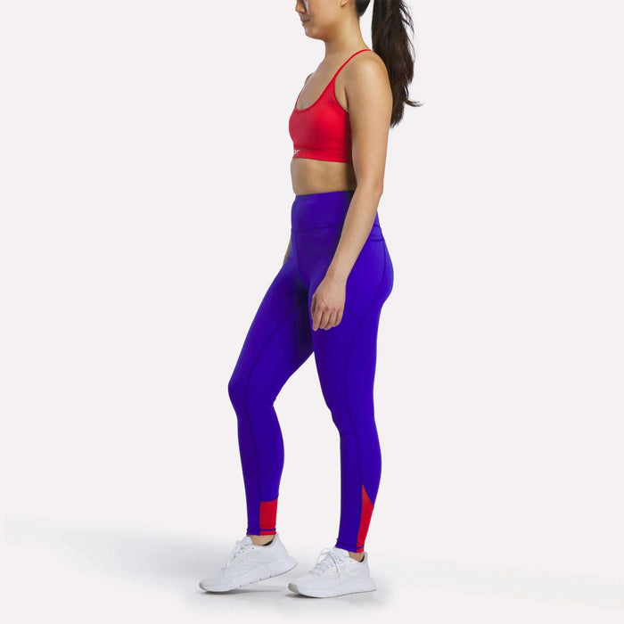 A woman stands sideways against a plain white background, hands relaxed by her sides, wearing a red sports bra and Reebok Women's Running Leggings in blue with red accents. The leggings feature stylish mesh panels for added breathability and are paired with white sneakers.