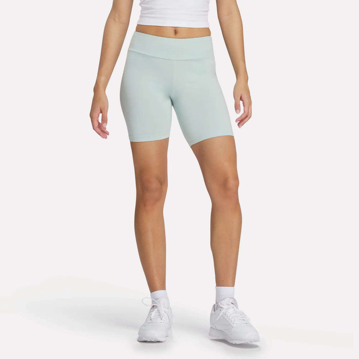 A person stands against a plain background wearing a white crop top, light blue Reebok Women's Small Logo Cycle Shorts with a stretchy fit, white socks, and women's Reebok athletic shoes. Their arms are relaxed at their sides.