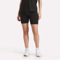 A person wears Reebok Women's Small Logo Cycle Shorts, a black T-shirt, and white sneakers against a plain white background. Only the lower half of the body is visible.